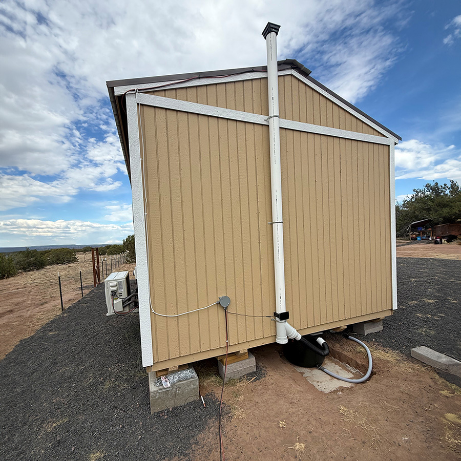A white vent pipe running up the outside wall of a small off-grid building to above the roofline, with the composting container below the floor