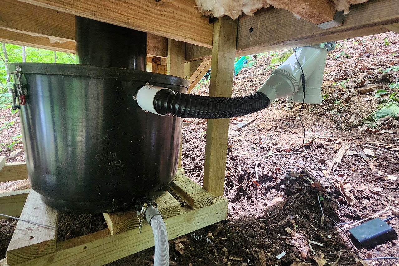 A black GL 90 composting container standing on timber under a cabin floor, with a ribbed vent hose and a liquid drain hose running from it
