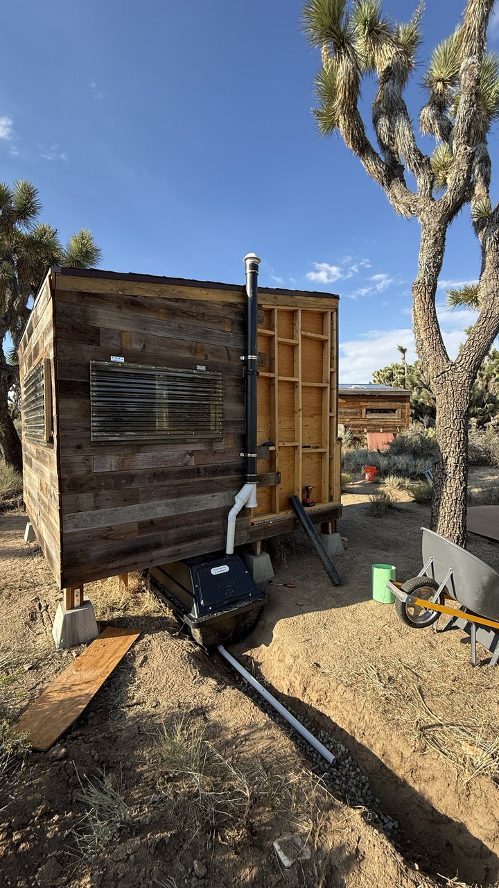 Rear of the outhouse: the CF 4 composting container below the floor, a black vent pipe running up the back wall past the roofline, and a white drain pipe in a gravel trench