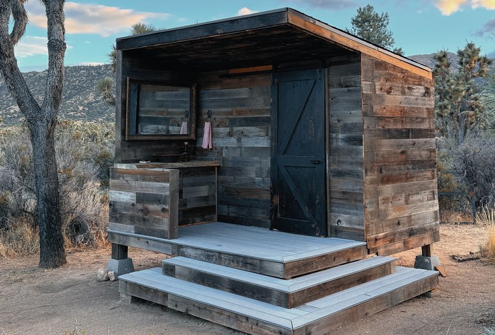 A modern reclaimed-wood outhouse with a black barn door and wide deck steps among Joshua trees