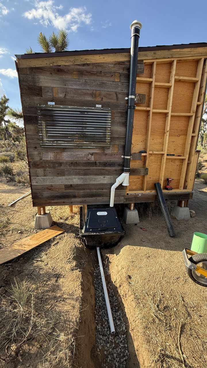 Close view of the CF 4 container's service hatch under the outhouse, with the vent pipe rising up the wall and the excess-liquid drain pipe laid in gravel