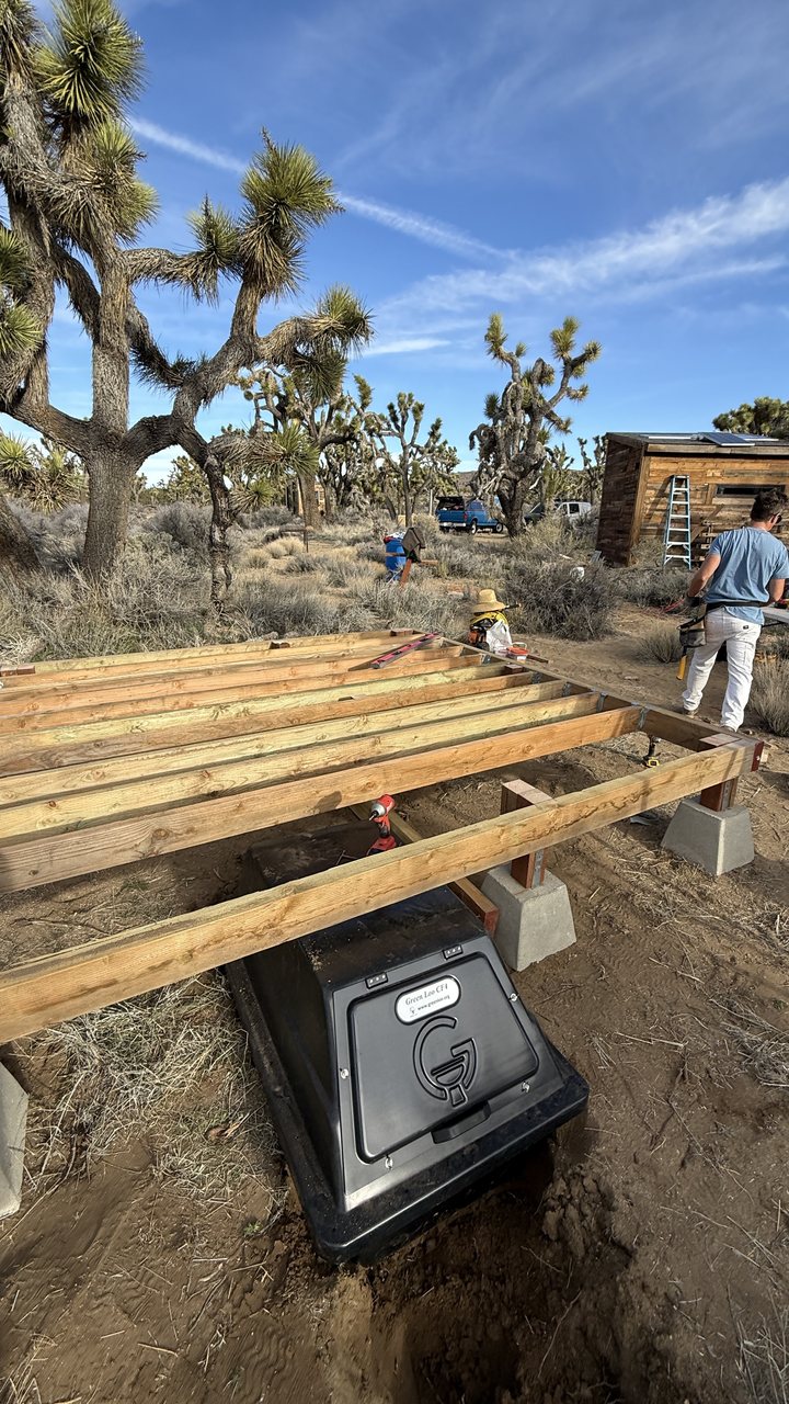 The black CF 4 composting container sitting in a shallow excavation directly under the outhouse floor joists