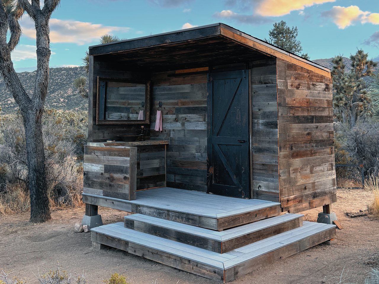 A modern reclaimed-wood outhouse with a black barn door, an outdoor vanity and wide deck steps, among Joshua trees with desert hills behind