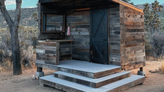 Front view of a reclaimed-wood outhouse with a black barn door, outdoor vanity and gray deck steps in the desert
