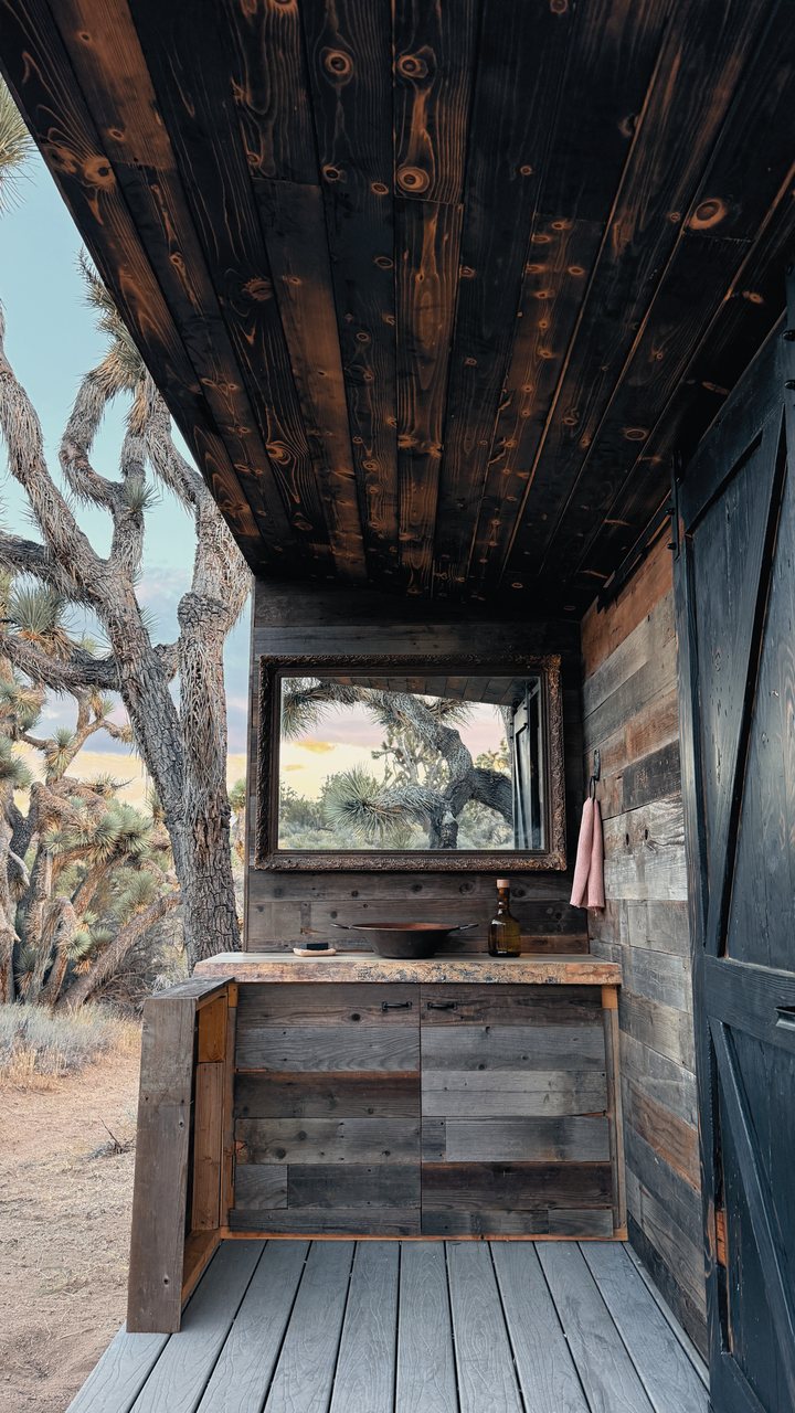 An outdoor vanity with a copper basin and an ornate framed mirror under a charred-wood ceiling, a Joshua tree reflected in the mirror