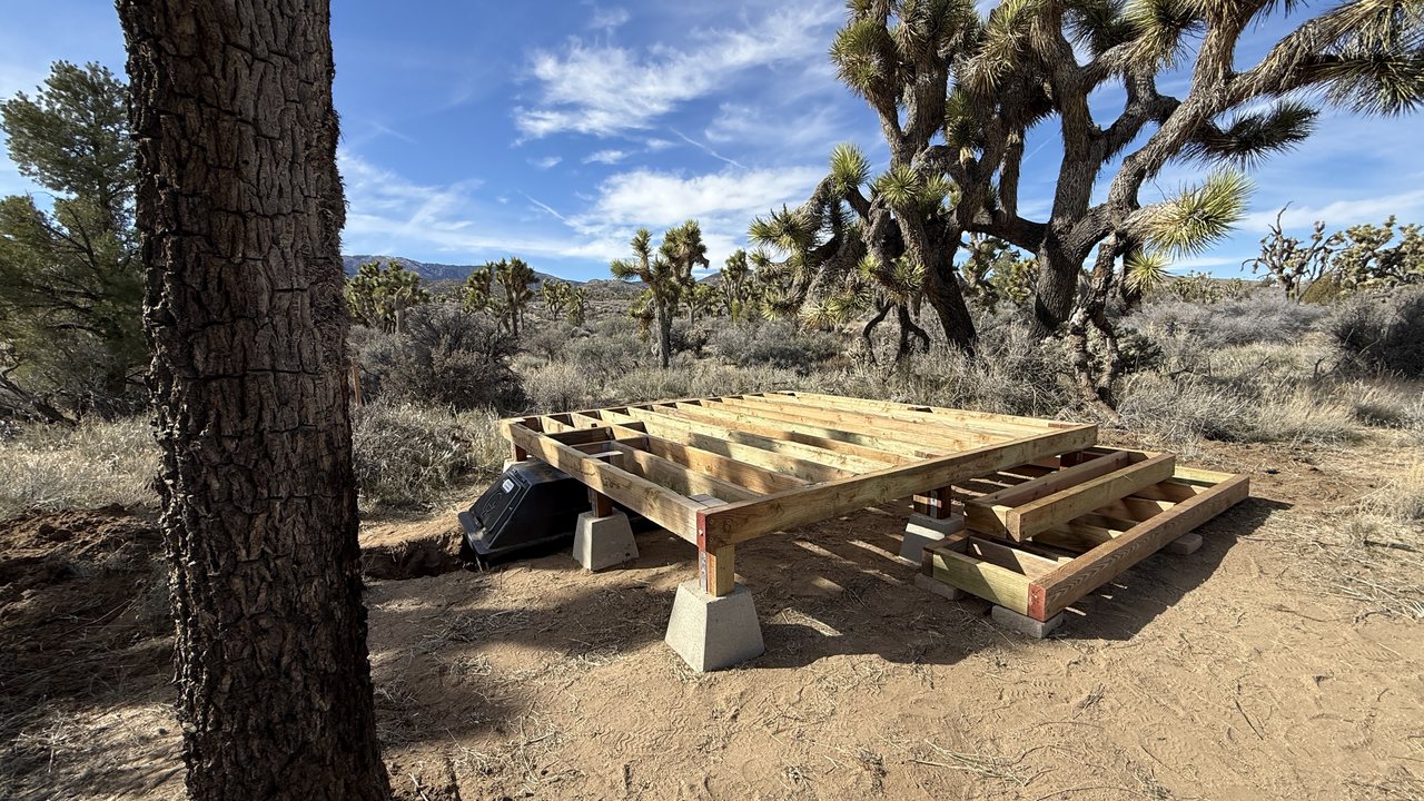 A timber floor frame on concrete pier blocks among Joshua trees, with the black CF 4 composting container already set in the ground beside it