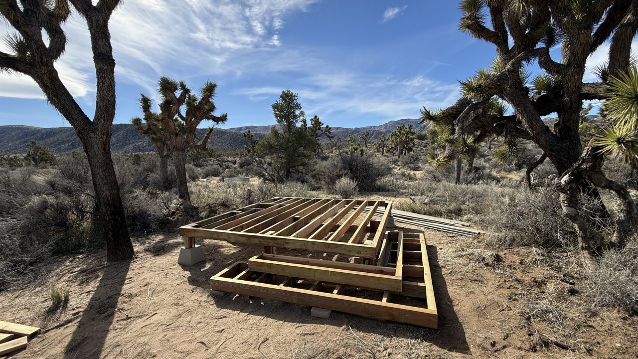 Two timber floor frames for an outhouse and its deck, laid out on the desert floor among Joshua trees with mountains behind