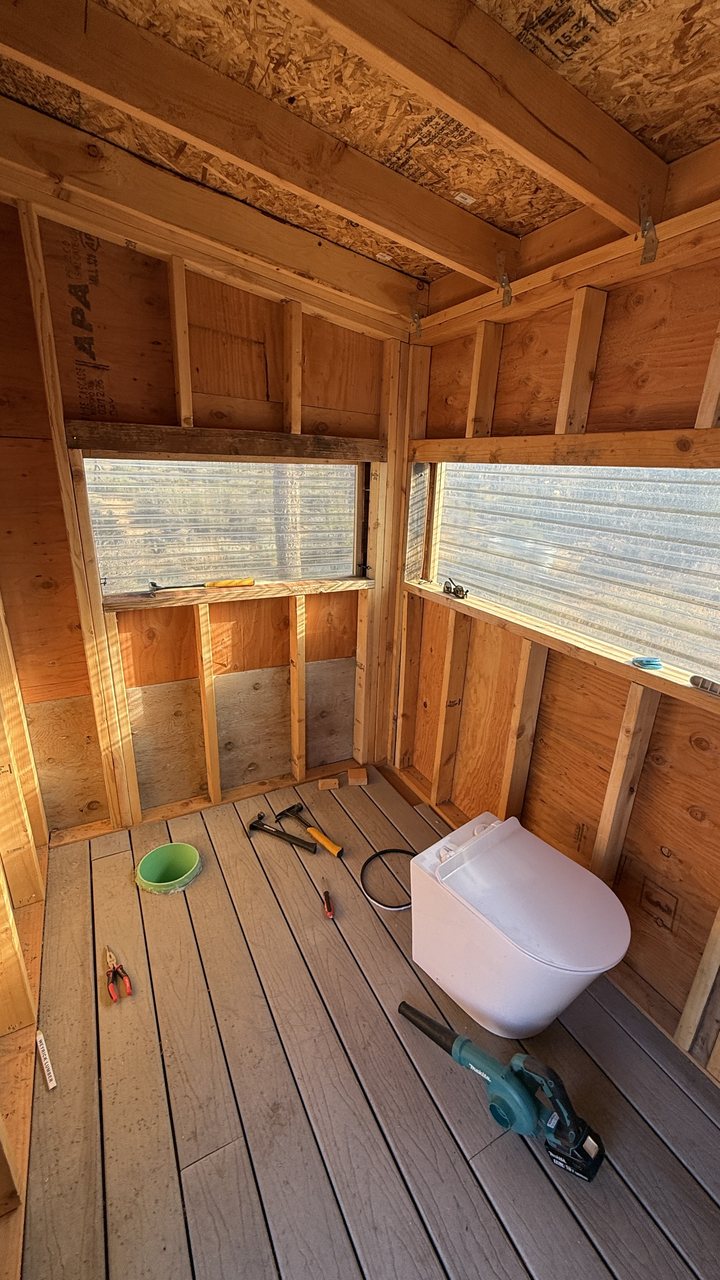 A white porcelain toilet pedestal installed on a composite deck floor inside the outhouse during construction, tools on the floor