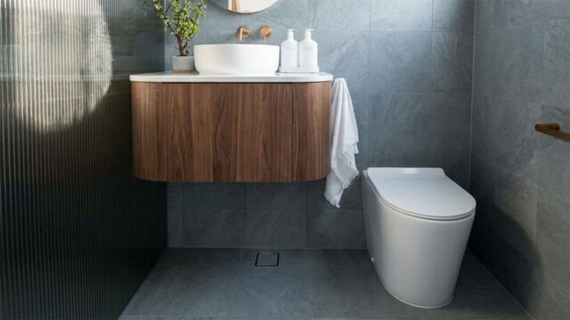 A white porcelain waterless toilet pedestal from Waterless Toilet Shop in a modern bathroom with grey stone-look tiles, a walnut wall-hung vanity, a round vessel basin and brass fixtures