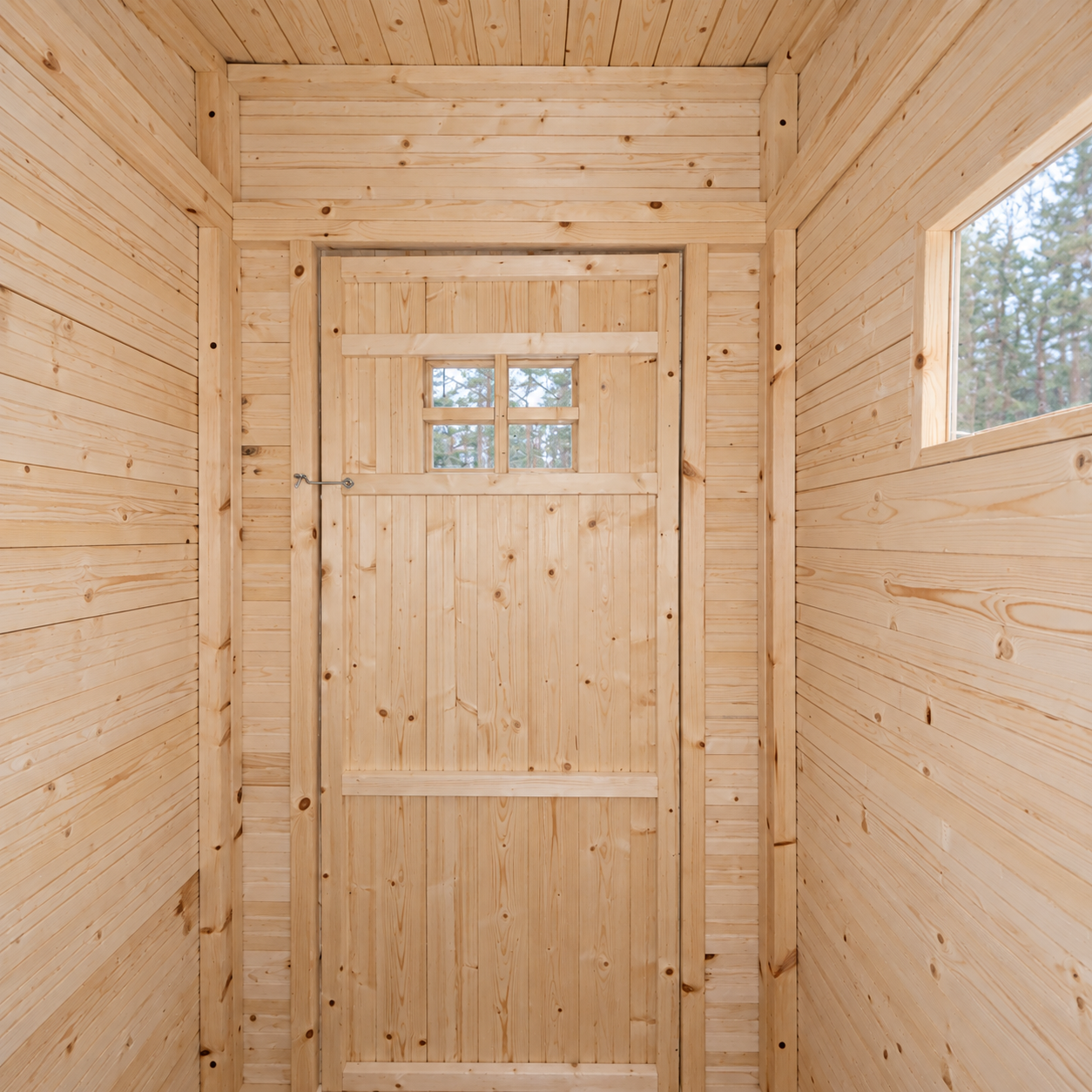 View from the seated position inside the pine Finnish Outhouse toward its closed four-pane window door, small metal privacy hook and single-pane side window.