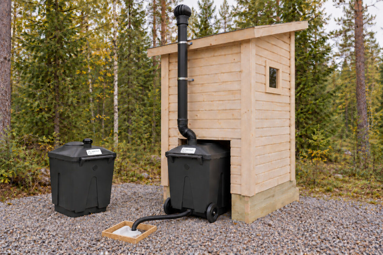 Rear view of the natural wood Finnish Outhouse with an installed black Green Toilet 330, a spare container, a ventilation pipe and a drain hose leading to a recessed liquid canister in a wooden enclosure.