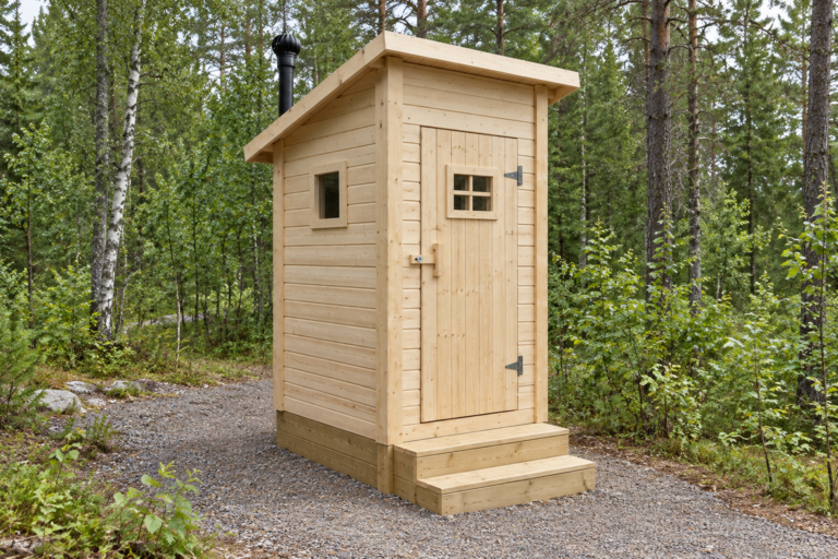 A small unpainted pine wooden outhouse with wooden steps on a gravel path in a birch and pine forest