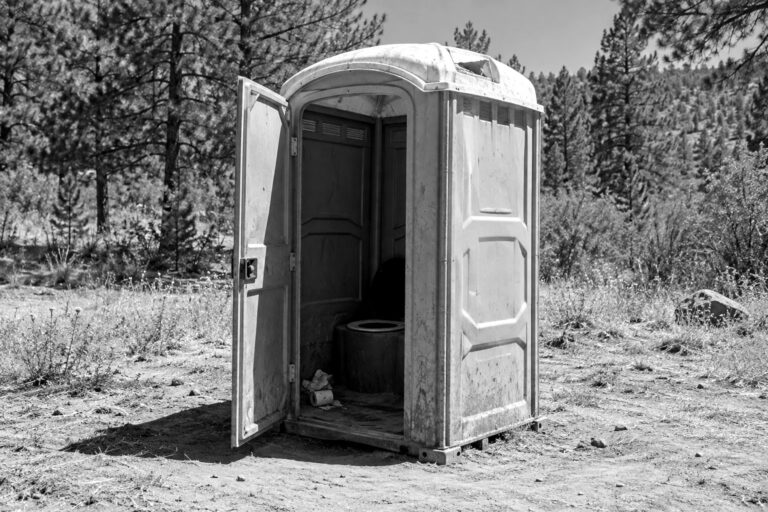 A dirty, sun-faded plastic portable toilet with its door half open on dry dirt at the edge of a campground, in black and white