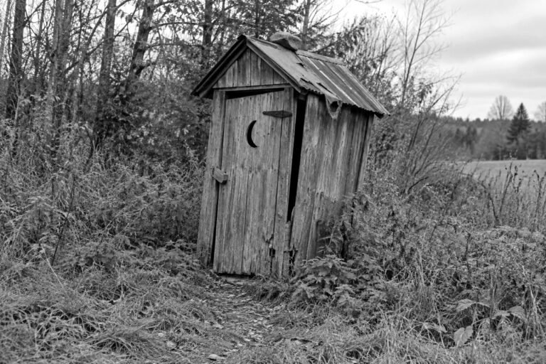 An old homemade wooden pit outhouse leaning to one side, with weathered boards, a crooked door and a tarp-patched roof, in black and white
